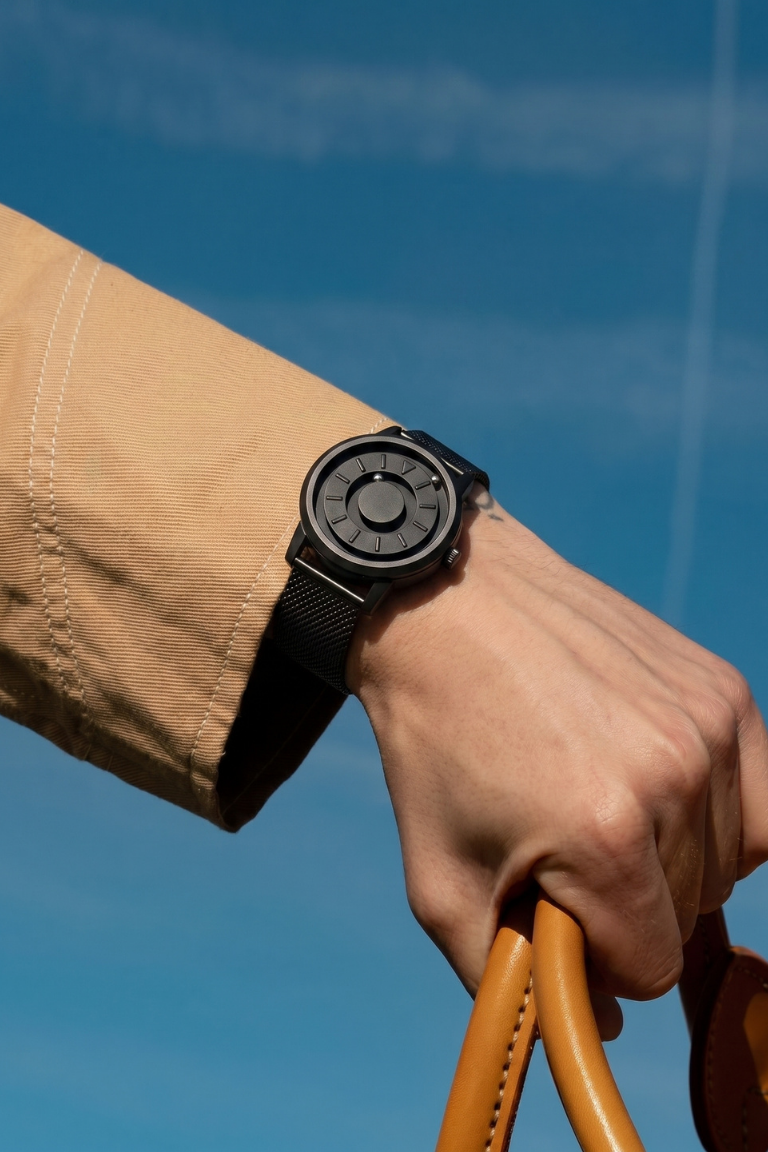 Hand wearing a black watch against a blue sky