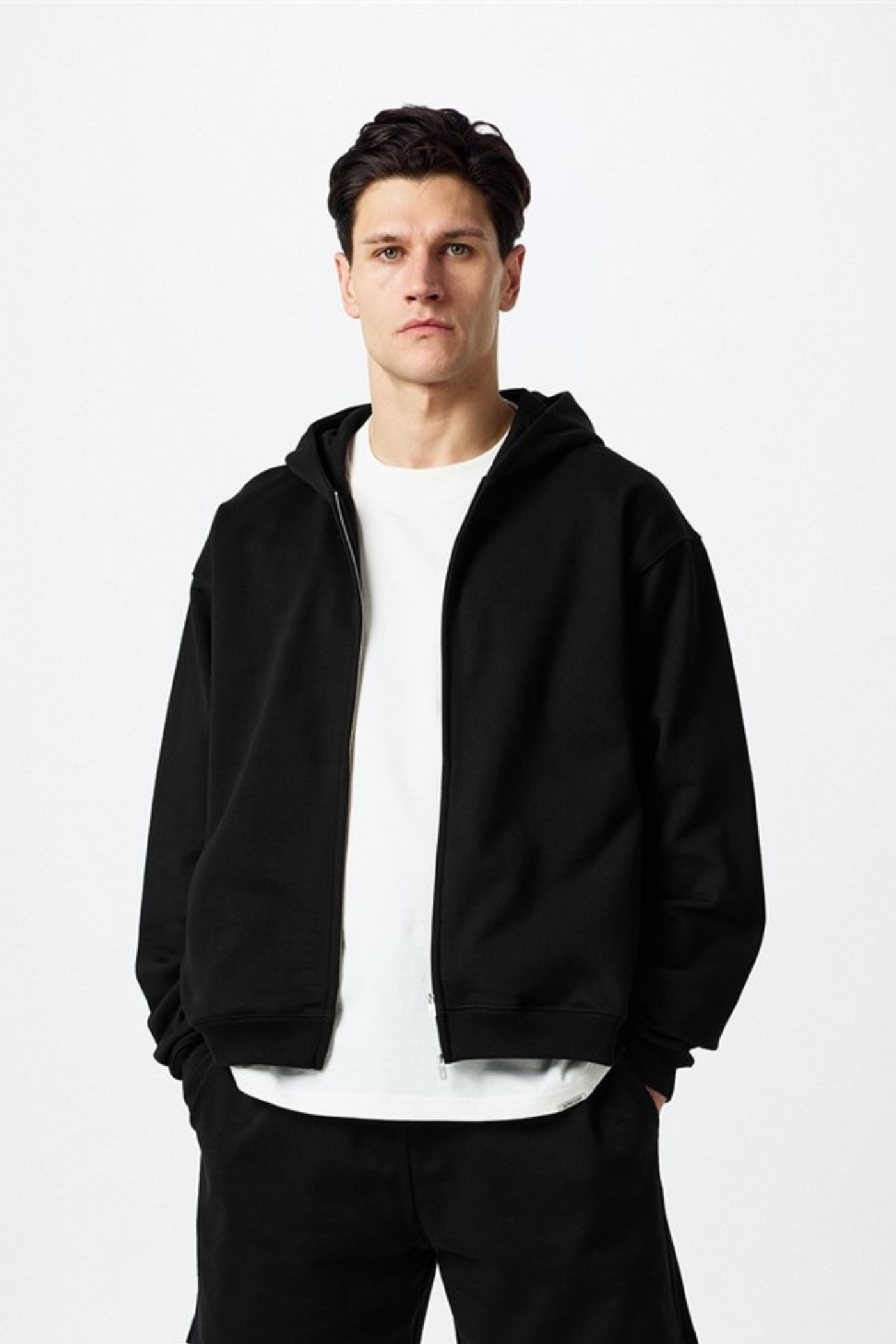 Man in black hoodie and matching pants over white t-shirt, standing against a white background.