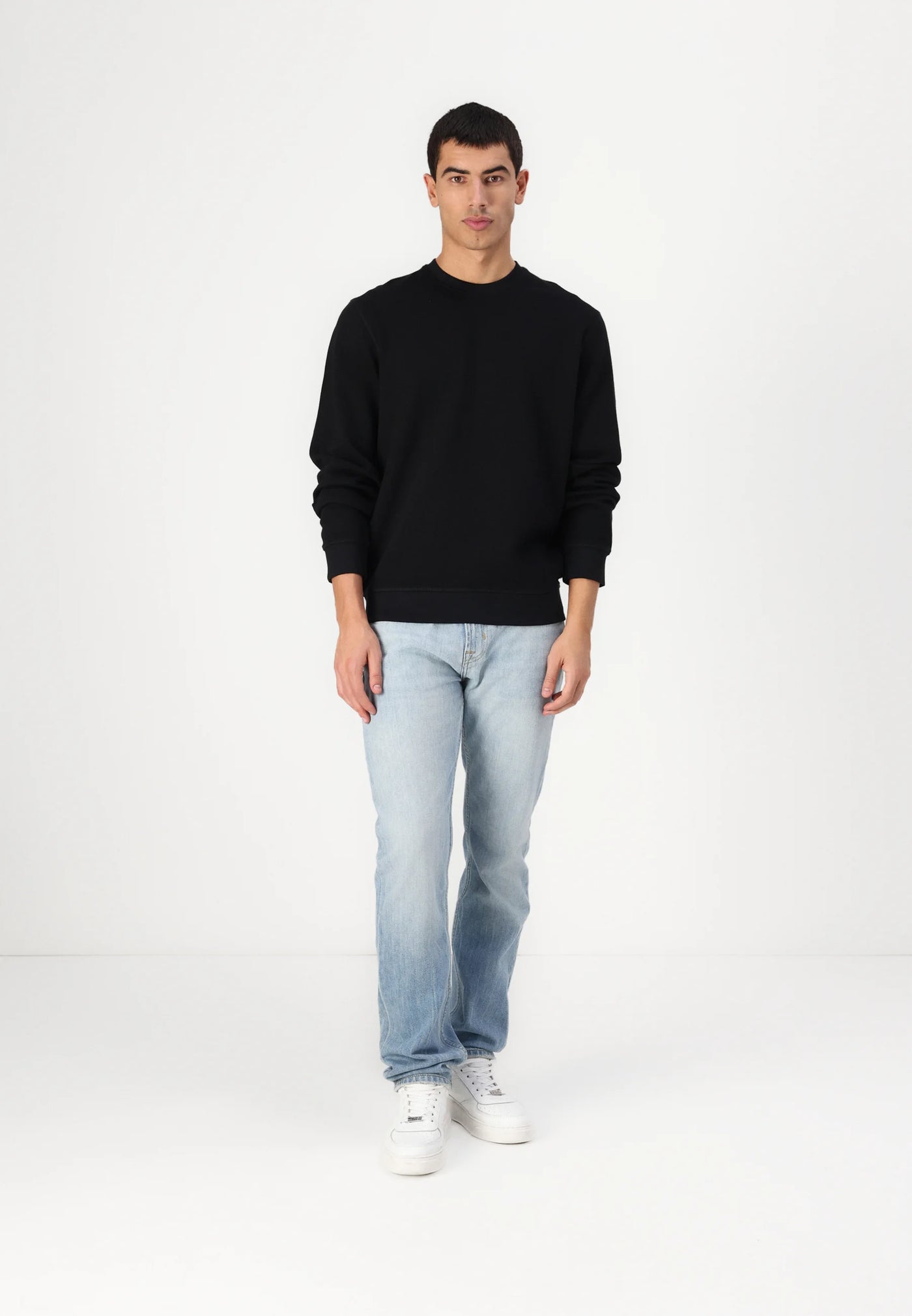 Man in black sweatshirt, jeans, and white sneakers stands in a white studio.
