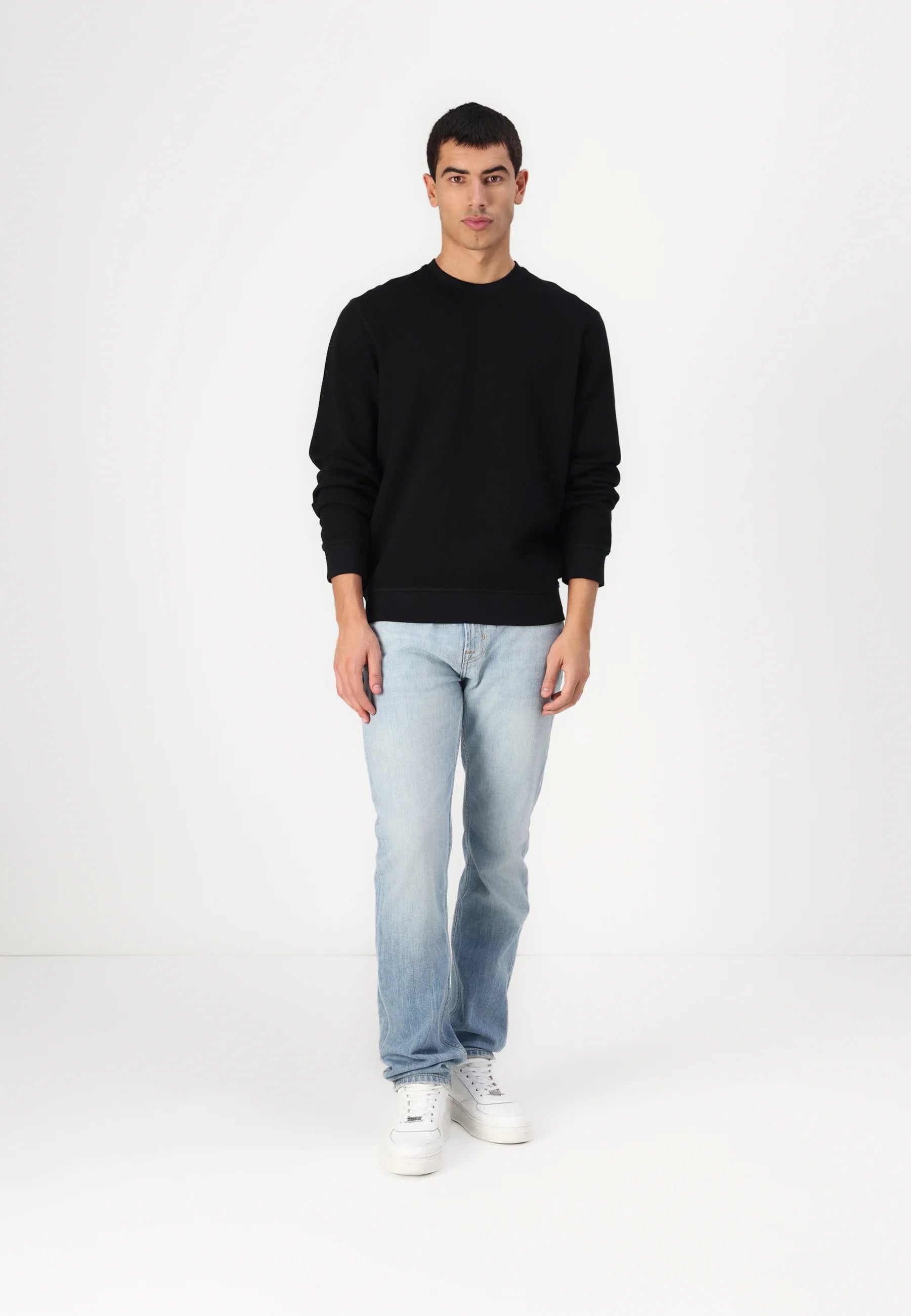 Man in black sweatshirt, jeans, and white sneakers stands in a white studio.